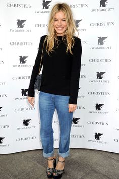 a woman standing in front of a white wall wearing blue jeans and black top with heels