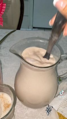 a person pouring milk into a glass pitcher on top of a table next to another cup