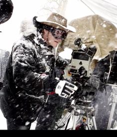 a man wearing a hat and glasses standing in the snow with a camera on a tripod