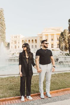 a man and woman standing next to each other in front of a fountain holding hands