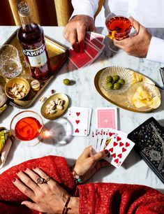 two people sitting at a table playing cards and drinking wine, with other items on the table