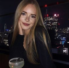 a woman sitting at a table with a drink in front of her and the city lights behind her