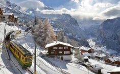 a yellow train traveling down tracks next to snow covered mountains and houses in the distance