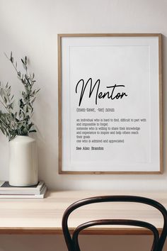 a white vase sitting on top of a wooden table next to a framed poster with the words mentor