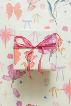 a gift wrapped in white paper with a pink ribbon on top of floral wallpaper