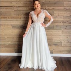 a woman standing in front of a wooden wall wearing a white dress with long sleeves