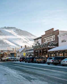 cars parked on the side of a road in front of buildings and snow covered mountains