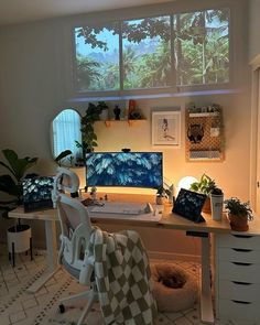 a desk with a computer on top of it in front of a window filled with potted plants