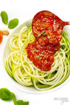 a white bowl filled with zucchini and tomato sauce on top of green leaves