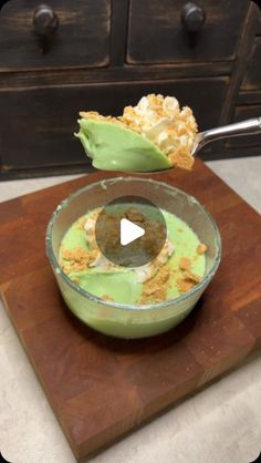 a wooden cutting board topped with a bowl filled with green liquid and toppings on top of it