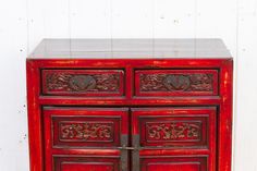 a red wooden cabinet with two doors and carvings on the front, sitting against a white wall