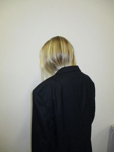 a woman with blonde hair standing in front of a white wall wearing a black jacket