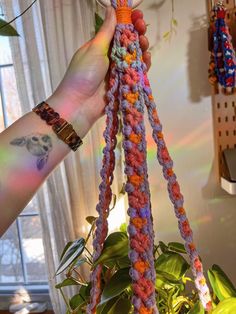 a person holding up a multicolored crochet plant hanger in front of a window