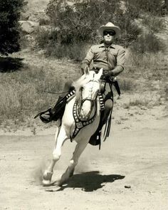 a man riding on the back of a white horse down a dirt road next to a forest