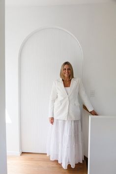 a woman standing in front of a white wall wearing a white jacket and skirt with her hands on her hips