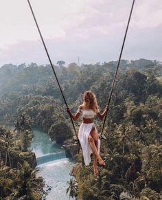 a woman is swinging on a rope in the air over trees and water with a waterfall behind her