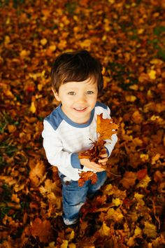 a young boy holding leaves in his hands while standing on the ground covered with autumn leaves