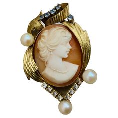 This is a 18K gold Cameo brooch/pendant. It depicts an oval shaped orange shell cameo with a relief of a woman bust. The woman has a wavy hair.She is wearing earrings. Her neck is adorned by a necklace. The cameo is mounted on a yellow gold bezel setting that is enhanced by a wreath made of engraved branches of leaves and a scroll of a ribbon. The wreath is embellished by five blue transparent tiny stones at the top, eight clear transparent tiny stones at the bottom and three pearls. The brooch is hallmarked with unrecognized marks at the needle of the safety catch. Cameo Brooch, How To Make Wreaths, Bezel Setting, Vintage Brooches, Wavy Hair, Jewelry Necklace Pendant, Vintage Jewelry, A Woman, 18k Gold