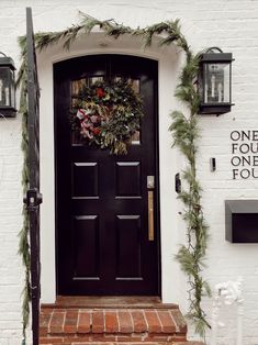 a black front door decorated with greenery and wreath on the side of a white brick building