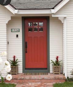 a red front door and brick walkway leading to a white house with flowers in the foreground