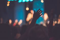 a person raising their hand up in front of a crowd at a music concert, with lights on the stage behind them