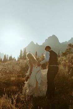 a bride and groom are walking through the grass in front of some tall mountains at sunset