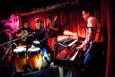 a group of men playing instruments in front of a keyboard and drums on stage with red curtains behind them