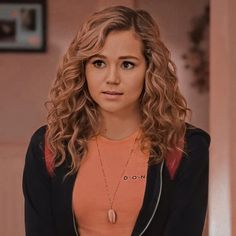 a woman with long curly hair wearing an orange shirt and black jacket is looking at the camera