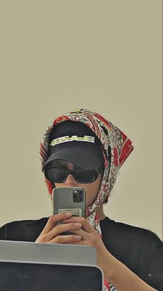 a woman wearing sunglasses and a headscarf is looking at her cell phone while sitting in front of a laptop