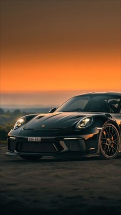 a black sports car parked on top of a dirt field next to the ocean at sunset