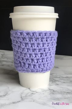 a crocheted coffee cup cozying in purple and white is sitting on a marble surface