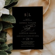 a black and white wedding card on top of some green leaves with flowers in the background