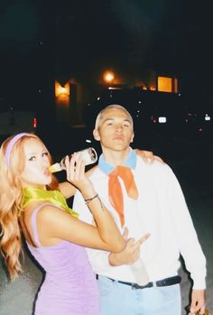 a man and woman standing next to each other drinking from bottles in the street at night