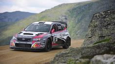 a rally car driving down a dirt road in front of some rocks and grass on the side of a mountain