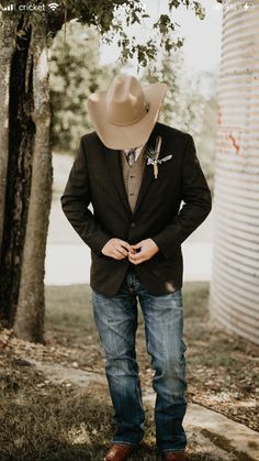 a man wearing a cowboy hat standing in front of a tree with his hands on his hips