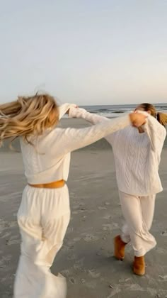 two women are dancing on the beach in white clothes and brown boots, with their hair blowing in the wind