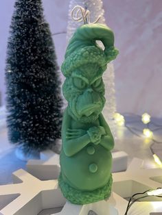 a green figurine sitting on top of a snow covered ground next to christmas trees