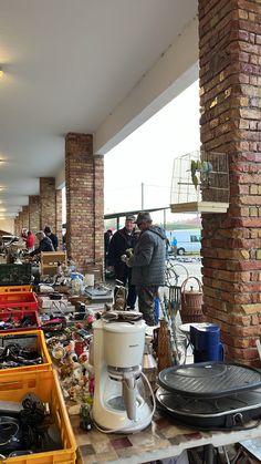 people looking at various items on a table in a room with brick pillars and windows