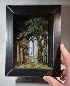 a hand holding up a miniature painting of a window in a forest with trees and grass