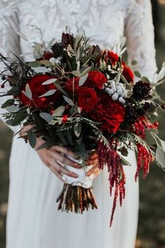 a woman in a white dress holding a red and black bouquet with greenery on it