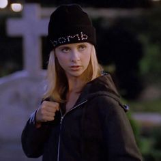 a woman wearing a black hat and jacket standing in front of a grave with her hand on her hip