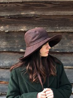 a woman wearing a brown hat standing in front of a wooden wall with her hands on her hips