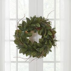 a wreath hanging on the side of a window in front of a white curtain with green leaves and pine cones