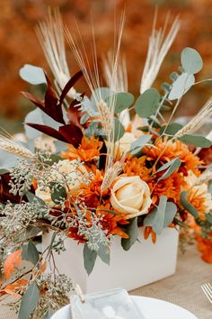 an arrangement of flowers and greenery in a square vase on a table with place settings