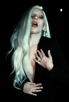 a woman with white hair and makeup is posing for the camera in black dress, holding her hands on her chest