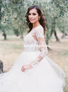 a woman in a white wedding dress standing under an olive tree with her hands on her hips