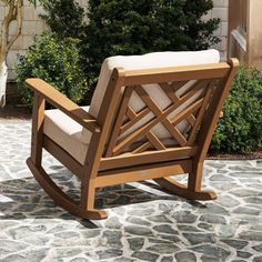 a wooden rocking chair sitting on top of a stone floor next to a tree and bushes