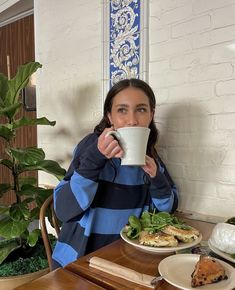 a woman sitting at a table drinking from a coffee cup with food on the side