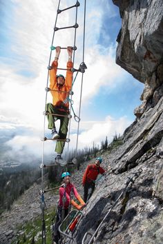 two people climbing up the side of a mountain with their hands in the air while another person is holding on to ropes