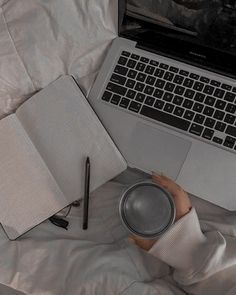 a person holding a coffee cup in front of a laptop on a bed with white sheets